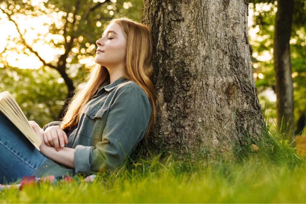 Young woman with long hair reading a book while sitting against a tree in a park.