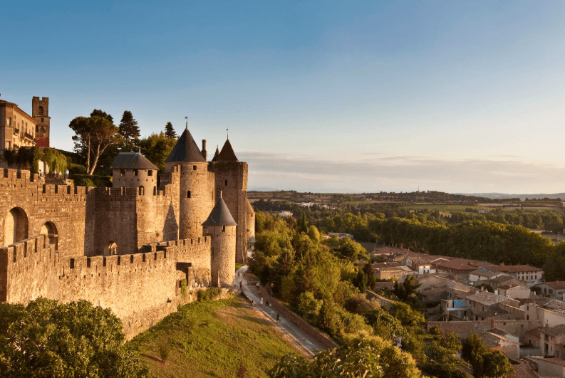 A medieval stone fortress with conical towers, part of the UNESCO World Heritage Sites in France, overlooks a small town and lush green landscape under a clear sky during sunset.