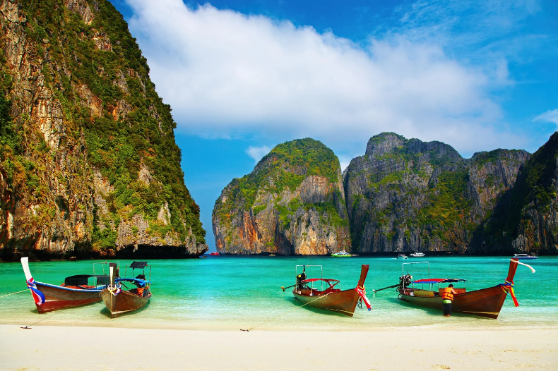 Three traditional wooden boats float on turquoise water near a white sandy beach—one of the prettiest beaches in the world—framed by dramatic green cliffs and rocky islands under a partly cloudy sky.