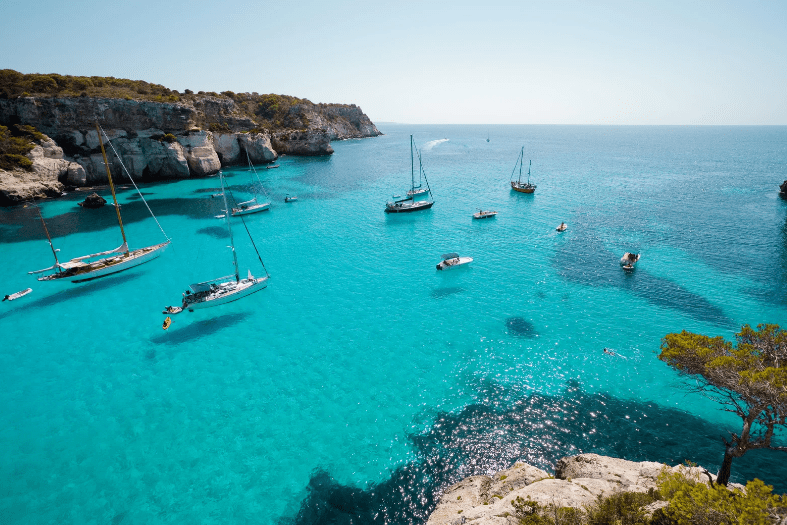 Several sailboats and small boats float on clear turquoise water near rocky cliffs with sparse greenery under a bright, sunny sky, capturing the charm of some of the prettiest beaches in the world.