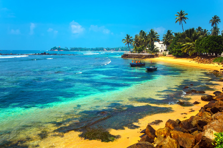 A tropical beach with golden sand, turquoise water, palm trees, and a few boats near the shore. White buildings and lush greenery line the coastline under a bright blue sky—one of the prettiest beaches in the world.