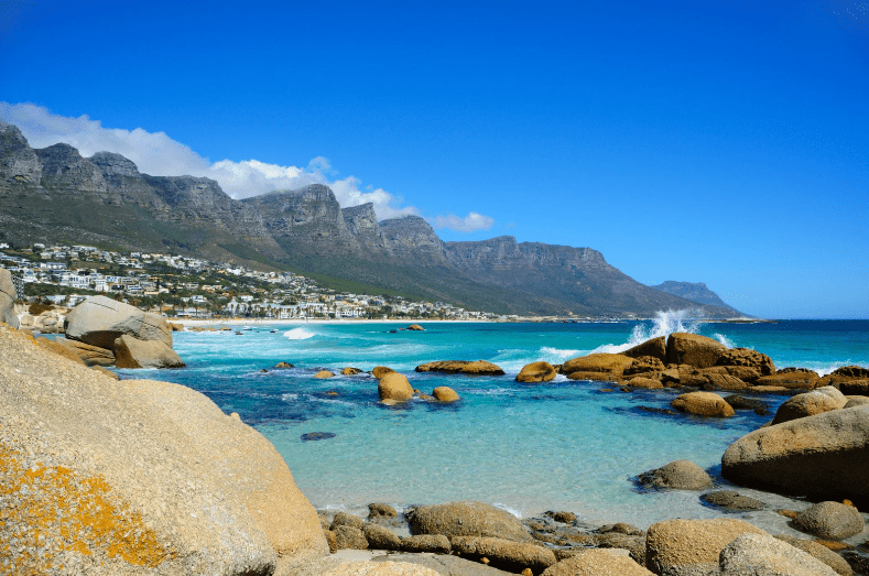 Clear blue ocean water with waves crashing on rocks, a sandy beach, and houses along the coastline—this spot stands among the prettiest beaches in the world. In the background, rugged mountains rise under a bright blue sky with scattered clouds.