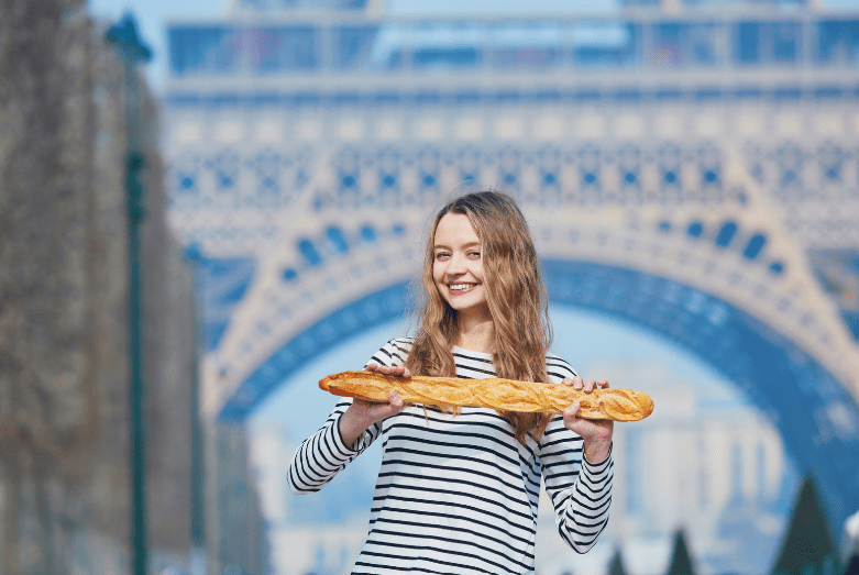 A smiling woman in a striped shirt holds a large baguette—a common food in France—in front of the Eiffel Tower in Paris, with trees and the iconic tower visible in the background.