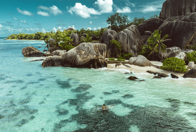 A person paddles a small kayak in clear turquoise water near one of the prettiest beaches in the world, surrounded by large granite boulders, palm trees, and lush greenery under a partly cloudy sky.
