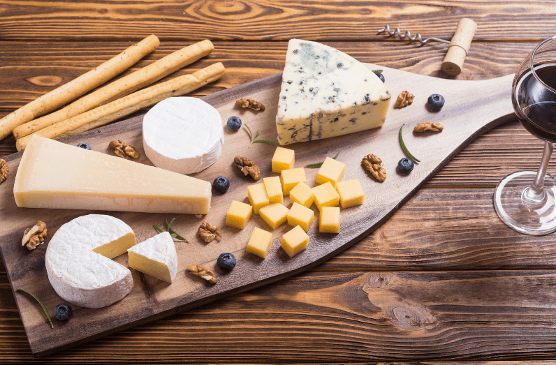 A wooden board with assorted cheeses—a common food in France—breadsticks, walnuts, blueberries, and a glass of red wine sits on a wooden table. The cheeses are in various shapes, including wedges, rounds, and cubes.