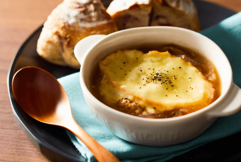 A bowl of French onion soup—a common food in France—topped with melted cheese and herbs, served on a plate with two pieces of rustic bread, a wooden spoon, and a turquoise napkin.
