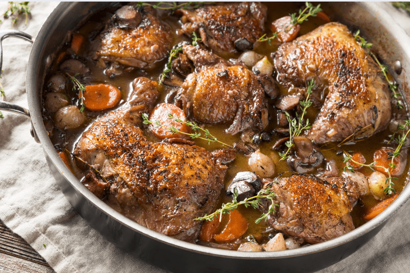 Common Food in France, A large pan filled with braised chicken thighs, carrots, pearl onions, mushrooms, and fresh herbs in a rich brown sauce—this common food in France is cooked and garnished with sprigs of fresh thyme.