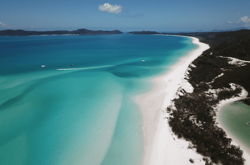 Aerial view of one of the prettiest beaches in the world, with turquoise water, white sand, and lush green vegetation lining the coastline beneath a clear blue sky.