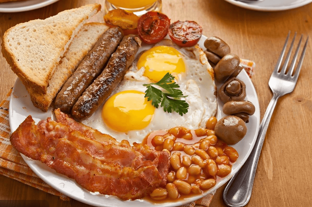 A plate of cooked breakfast, a common food in England, with sunny-side-up eggs, bacon, sausages, baked beans, grilled tomato, mushrooms, and toast garnished with parsley. A fork and a glass of orange juice are nearby.