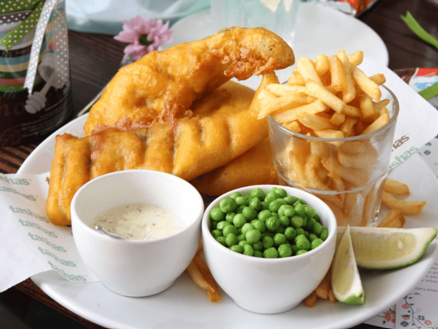 A plate of golden battered fish, a glass filled with French fries, a small bowl of green peas, tartar sauce, and a wedge of lime—this common food in England is arranged neatly on a white plate.