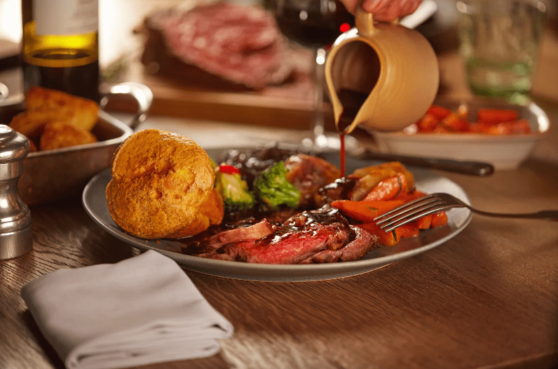 A plate of roast beef, Yorkshire pudding, roasted carrots, broccoli, and potatoes—a common food in England—is on the table. Someone is pouring gravy over the meal. A napkin, wine bottle, and extra food are in the background.