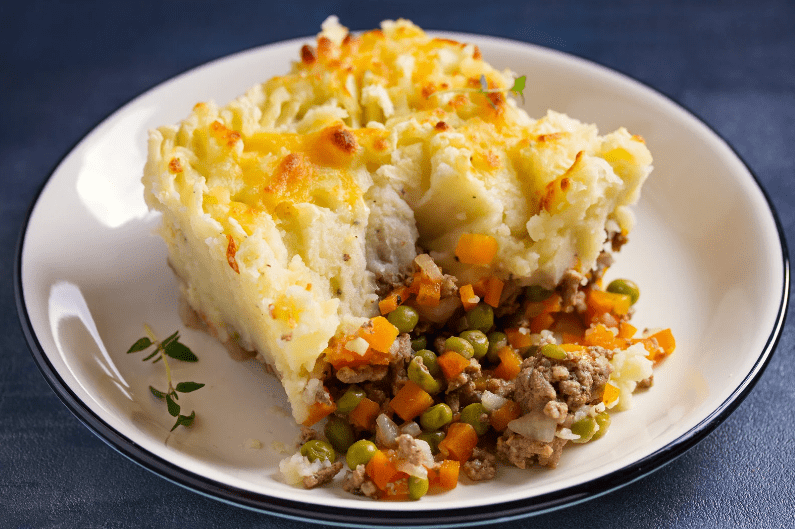 A serving of shepherd’s pie on a white plate, showing a layer of mashed potatoes on top and a filling of ground meat, peas, carrots, and onions underneath. A sprig of fresh thyme is on the side—a classic common food in England.