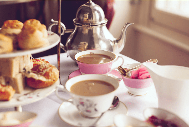 A close-up of a table set for afternoon tea, featuring pink and white china cups filled with tea—a common food in England—alongside a silver teapot, a tiered tray with scones and pastries, and a bowl with pink napkins.