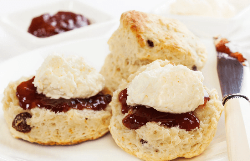 Two scones on a plate, one split and topped with red jam and a dollop of clotted cream—a common food in England. A knife with jam and more scones and cream are visible in the background.