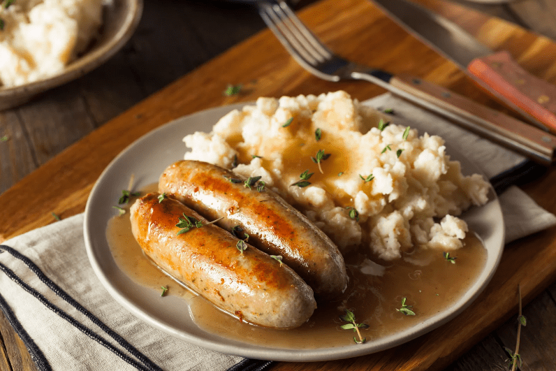 A plate with two grilled sausages topped with gravy, served alongside creamy mashed potatoes and garnished with fresh herbs—a classic common food in England. A fork and knife rest nearby on a wooden table.