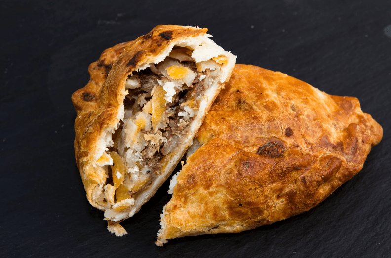 A golden-brown pasty with a flaky crust is cut open, revealing a savory filling of meat, potatoes, and vegetables—a delicious example of common food in England—set against a dark slate background.