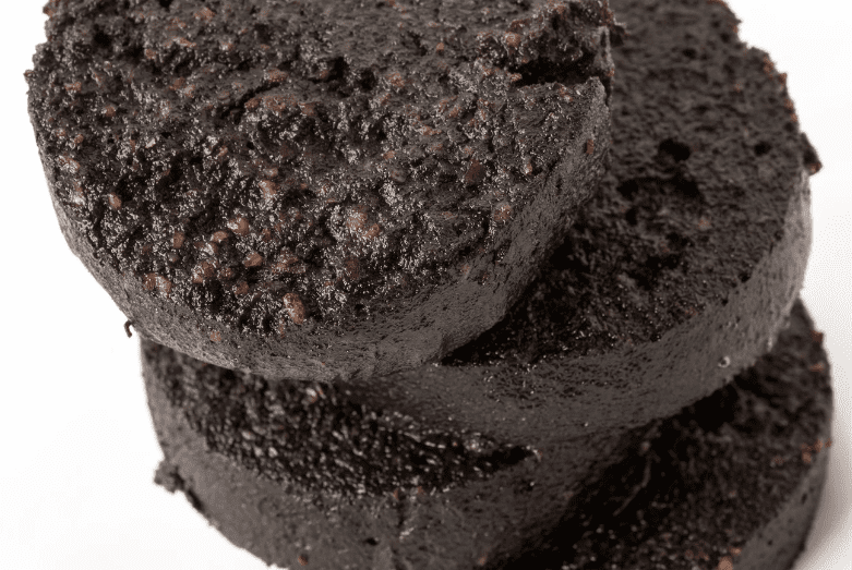 Close-up of three thick, round slices of black pudding stacked on top of each other, showing a moist, dark, crumbly texture with visible grains and fat—a common food in England.