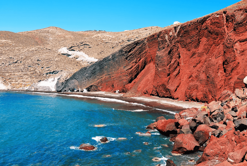 A vivid red volcanic cliff borders a small sandy beach beside turquoise blue water, with dry hills in the background under a clear blue sky—one of the prettiest beaches in the world.