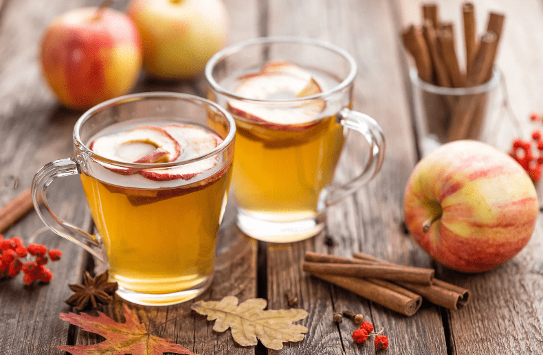 Two glass mugs of apple cider, a common food in England, with apple slices sit on a wooden table surrounded by apples, cinnamon sticks, star anise, red berries, and an autumn leaf, creating a cozy fall scene.