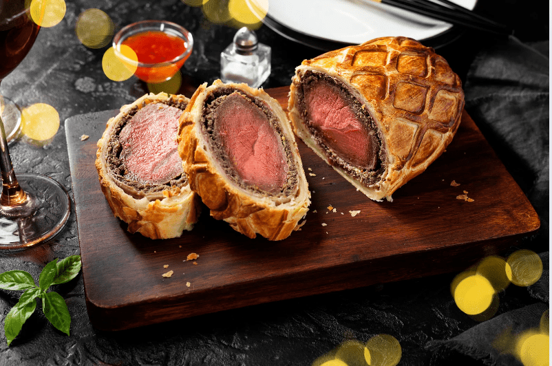 Two slices of beef Wellington, a common food in England, with golden, flaky pastry and a pink beef center sit on a wooden board. A glass of red wine, sauce, and basil garnish are nearby, with festive yellow lights in the background.