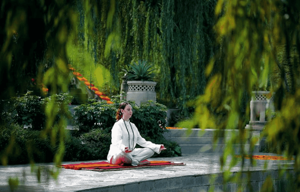 A woman in white clothing sits cross-legged on a mat, meditating outdoors in a lush, green garden—capturing the peaceful essence of meditation retreats in India, surrounded by leafy plants and stone steps.