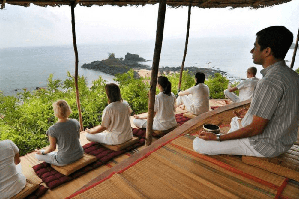A group of people sit cross-legged on mats under a thatched roof, participating in one of the serene meditation retreats in India, meditating as they overlook a scenic ocean view with a rocky shoreline and lush greenery.