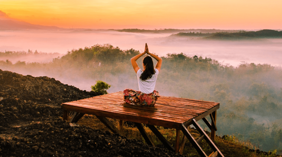 Meditation retreats in India, A person sits cross-legged on a wooden platform, facing a misty sunrise landscape with hands raised in a yoga pose—capturing the peaceful, serene atmosphere often found at meditation retreats in India.