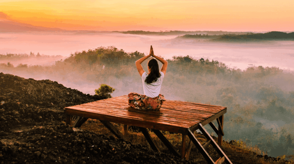 A person sits cross-legged on a wooden platform, practicing yoga at sunrise, facing misty hills and a colorful sky—an atmosphere reminiscent of meditation retreats in India.
