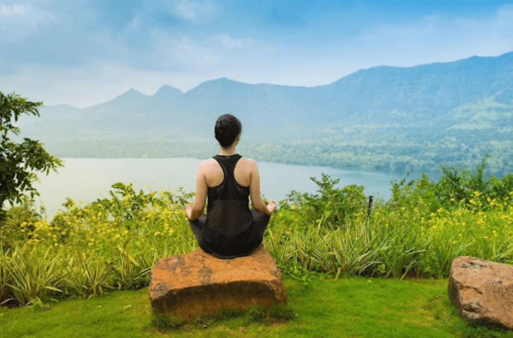 A person sits cross-legged on a stone, meditating and facing a scenic view of a lake, lush greenery, and distant mountains under a blue sky—reminiscent of tranquil meditation retreats in India.