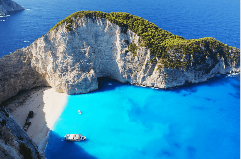 Aerial view of Navagio Beach on Zakynthos Island, Greece—one of the prettiest beaches in the world—featuring turquoise waters, a white sand shore, steep cliffs, lush greenery, and boats near the beach.