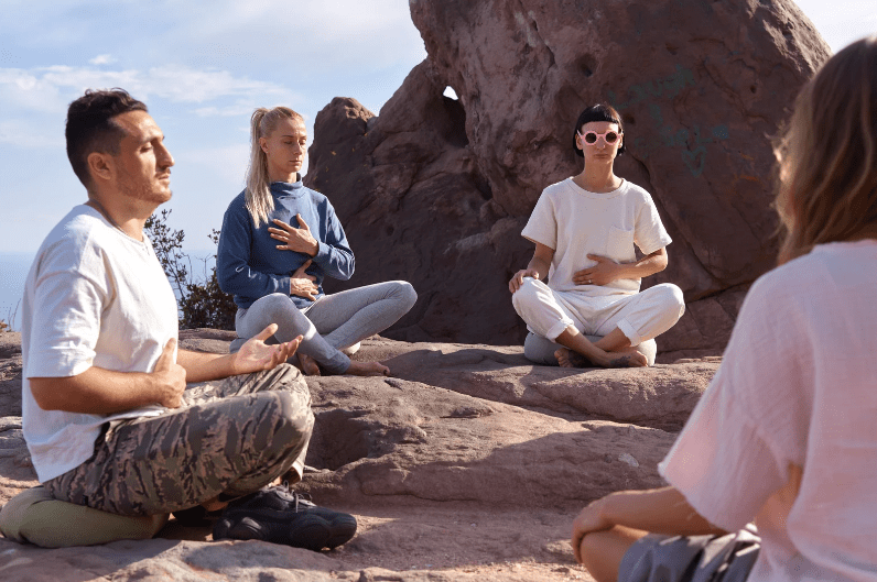 Four people sit cross-legged on rocky ground outdoors, meditating together as in meditation retreats in India. One person wears pink sunglasses, and all appear calm and focused, with a large rock formation behind them under a blue sky.
