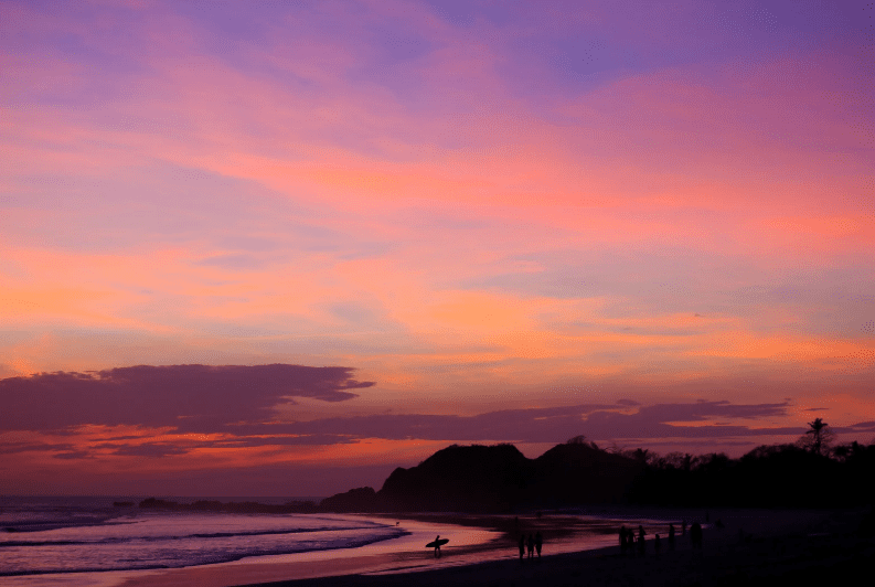 A beach at sunset with vibrant pink, purple, and orange clouds in the sky, silhouettes of hills and trees, people, a surfer on the shoreline—an idyllic scene reminiscent of Yoga Retreats in Costa Rica.
