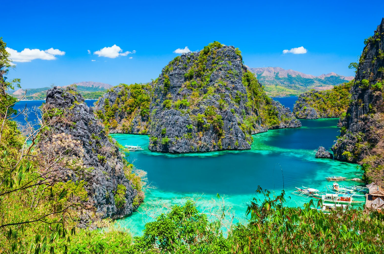Turquoise water surrounds limestone cliffs and lush green vegetation, with several small boats floating near the shore under a clear blue sky—an idyllic scene found on some of the prettiest beaches in the world.