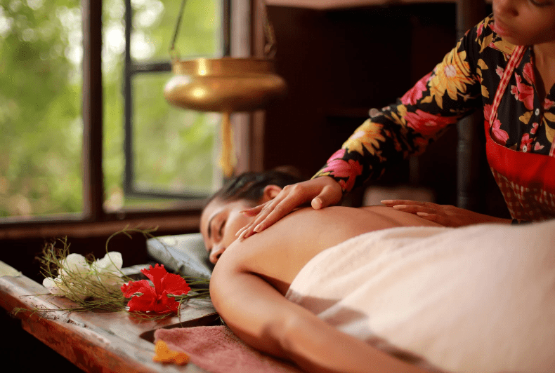 A person receives a relaxing back massage in a spa setting, reminiscent of yoga retreats in Costa Rica, lying face down on a massage table with a towel covering their lower body. Decorative flowers and soft natural light fill the tranquil room.
