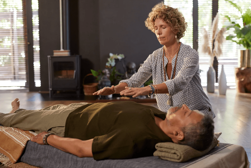 A woman performs a healing or energy therapy session on a man lying down with his eyes closed, indoors on a mat. Soft natural light and plants set a tranquil scene, reminiscent of the serenity found at Yoga Retreats in Costa Rica.