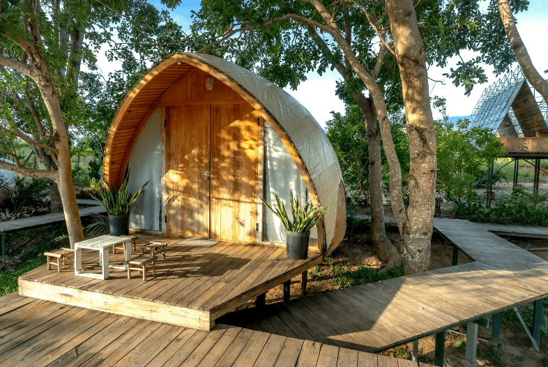 A small, arched wooden cabin with a front deck, surrounded by trees—perfect for Yoga Retreats in Costa Rica. Two potted plants, a table, and chairs sit on the deck. Wooden walkways connect the cabin to other areas amidst lush greenery.