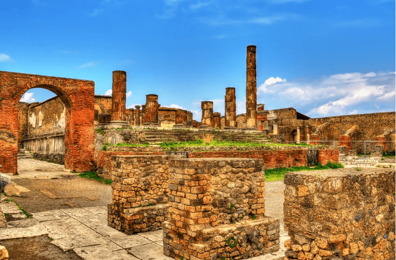 Ancient ruins with stone columns, arches, and weathered brick walls under a bright blue sky, these remnants surrounded by grassy patches evoke the grandeur of UNESCO World Heritage Sites in Italy.