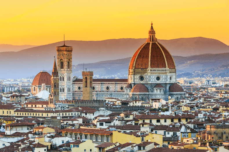 Florence Cathedral with its red-tiled dome and bell tower rises above the rooftops of Florence, Italy—one of the stunning UNESCO World Heritage Sites in Italy—set against a backdrop of hills at sunset.
