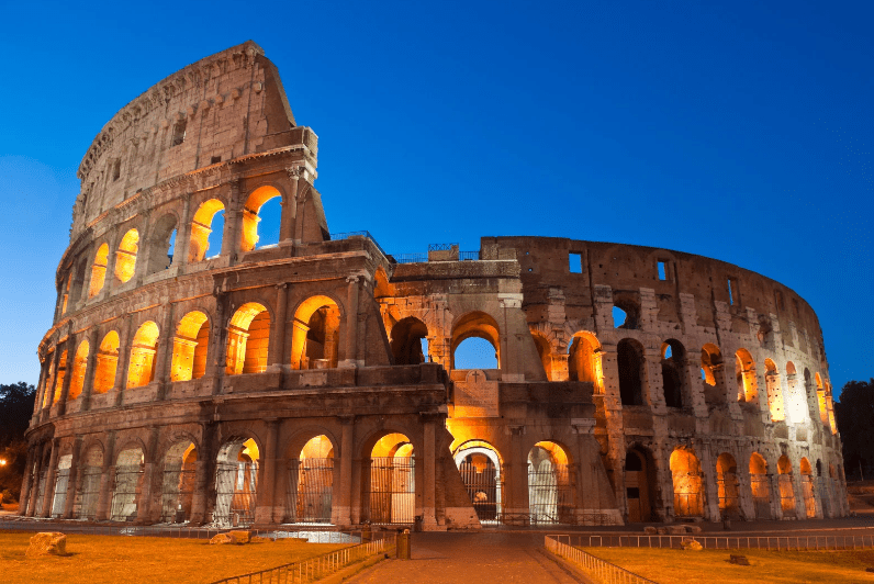 The Colosseum in Rome, Italy, a highlight among UNESCO World Heritage Sites in Italy, is illuminated with warm lights at dusk, showcasing its ancient stone arches and partially ruined structure against a clear blue sky.