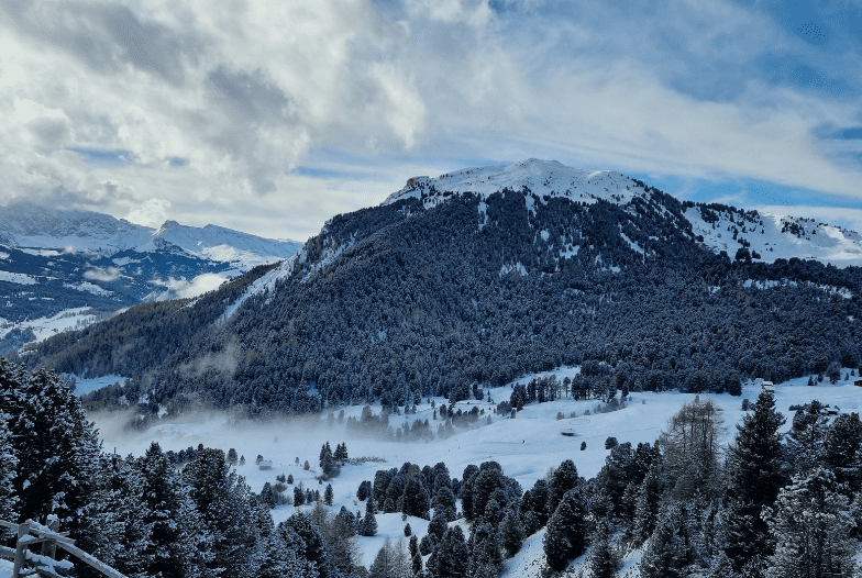 Snow-covered mountains and hills with dense evergreen forests under a partly cloudy blue sky evoke the tranquil beauty of UNESCO World Heritage Sites in Italy. Mist rises from the lower slopes as patches of snow blanket this serene winter landscape.