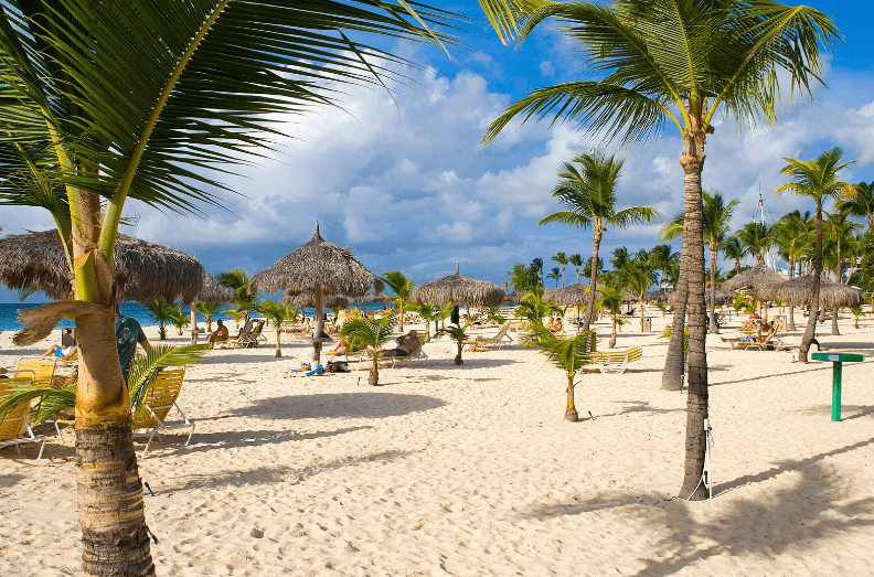 A sunny tropical beach with white sand, palm trees, thatched umbrellas, and lounge chairs. People relax under the shade, enjoying the bright blue sky and scattered clouds on one of the prettiest beaches in the world.