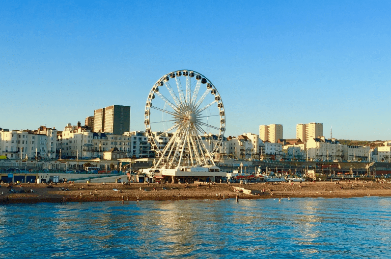 A large Ferris wheel stands near the shoreline, with city buildings and apartments behind it under a clear blue sky. The beach and waterfront are visible in the foreground, making this one of the hidden gems in England to discover.