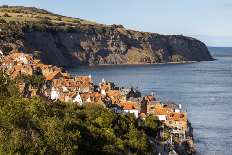 A coastal village with red-roofed houses nestled among green hills, beside calm blue water and steep cliffs under a clear sky. One of the Hidden Gems in England, people walk along a path in the foreground.