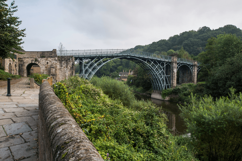 An old iron bridge with intricate metalwork spans a green, tree-lined river valley—one of the hidden gems in England—with stone walls, a historic building, and a cobbled walkway in the foreground beneath a cloudy sky.