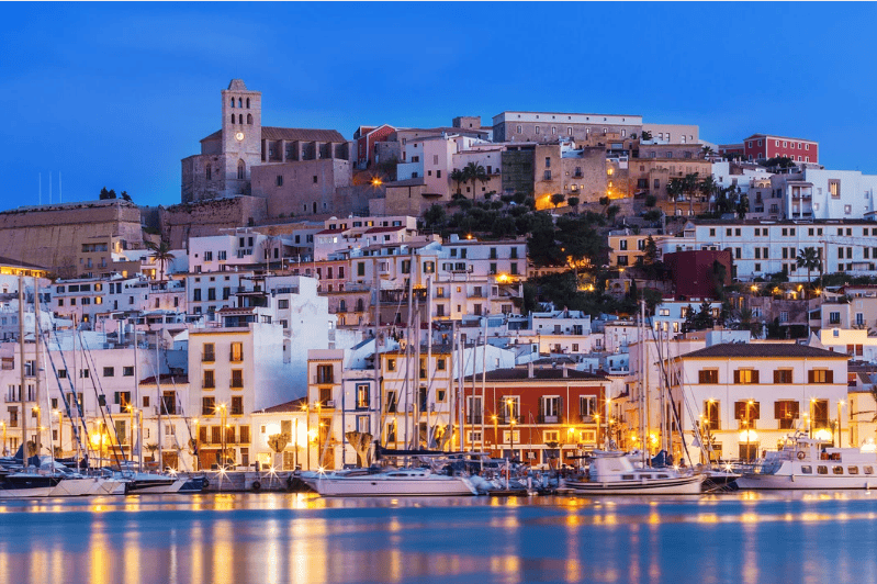 Tourist Attractions in Spain, View of Ibiza Town with illuminated buildings and boats in the harbor during dusk.