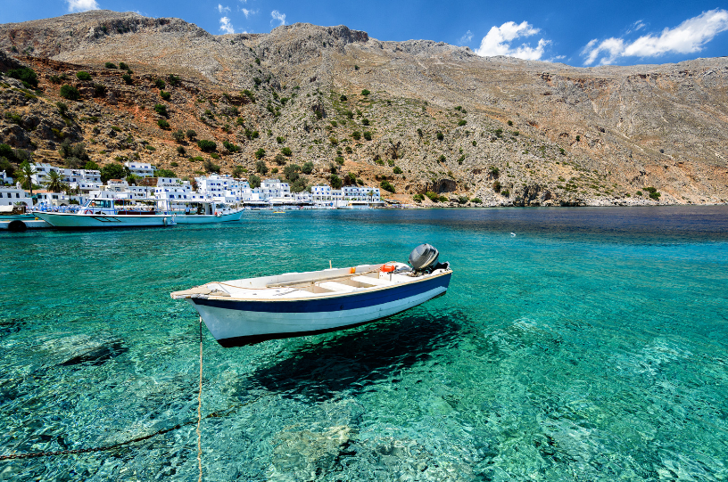 Turquoise harbor in Santorini with a white boat anchored near a rocky shoreline and whitewashed buildings along the coast of Greece. Tourist Attractions in Greece