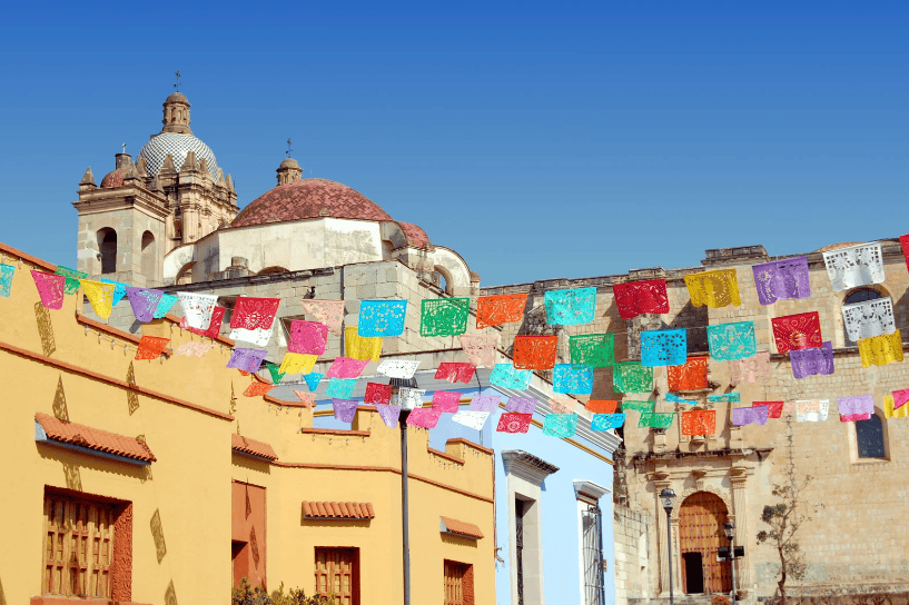 Colorful papel picado banners strung across buildings with a church dome in the background in a clear blue sky.Hidden Gems in Mexico